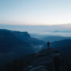 Silhouette of a lone hiker standing on a rocky outcrop above mist-filled blue valleys at sunrise, blue mountains decepcion